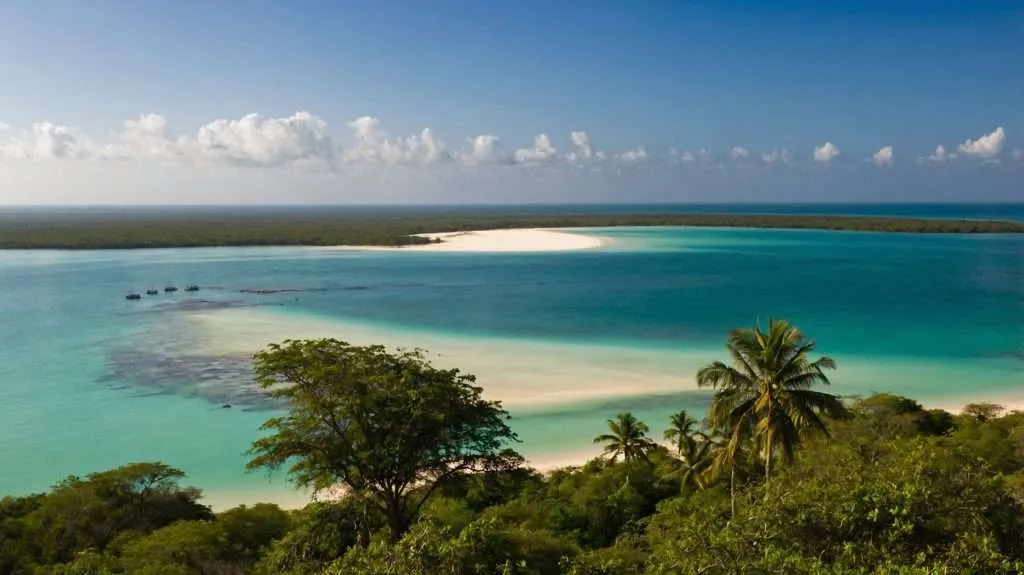 A scenic coastal view in Mozambique featuring turquoise waters, a white sandbar, palm trees, and anchored boats under a clear blue sky.
