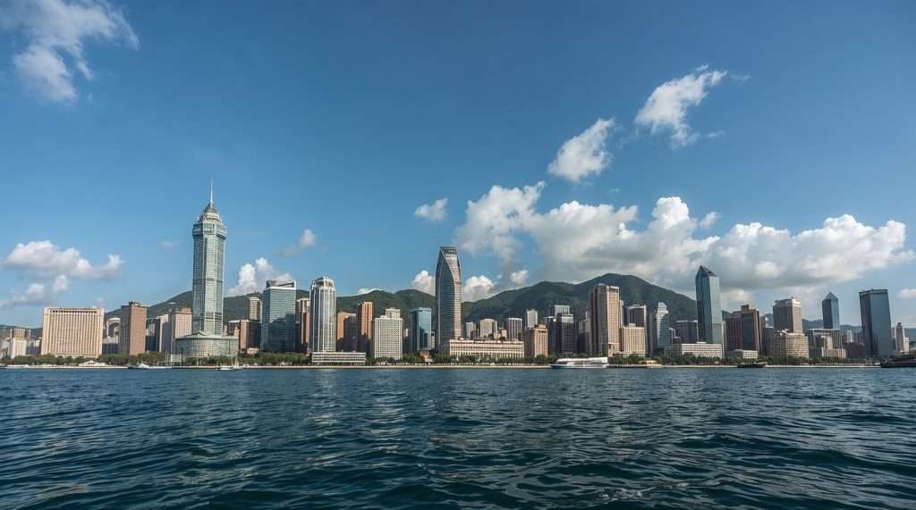 View of Macau's skyline from the sea, showcasing a range of modern skyscrapers against a backdrop of green hills under a clear blue sky with scattered clouds.