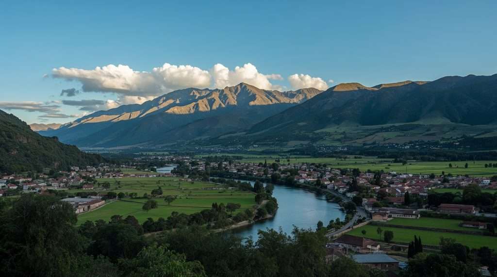 Scenic view of a Peruvian landscape featuring a winding river, green fields, and a village nestled below majestic mountains under a clear blue sky with scattered clouds.