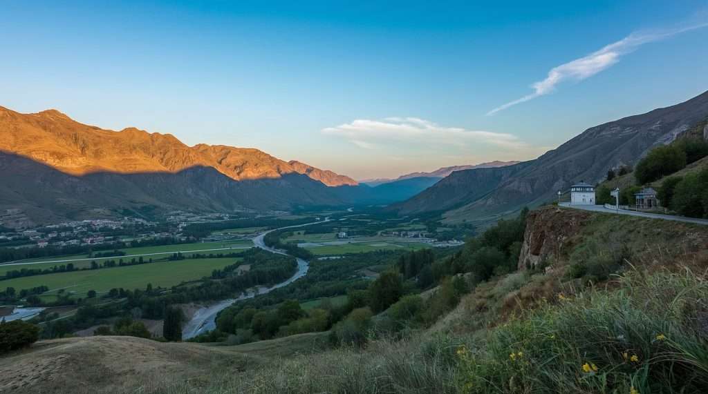 A scenic view of Azerbaijan’s landscape featuring a lush green valley, winding river, and distant mountains under a clear blue sky. A small building is perched on a hillside.