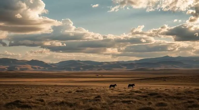 Vast Mongolian landscape under a dramatic sky with large clouds. Rolling hills stretch across the horizon, while two horses graze on the open grasslands in the foreground.