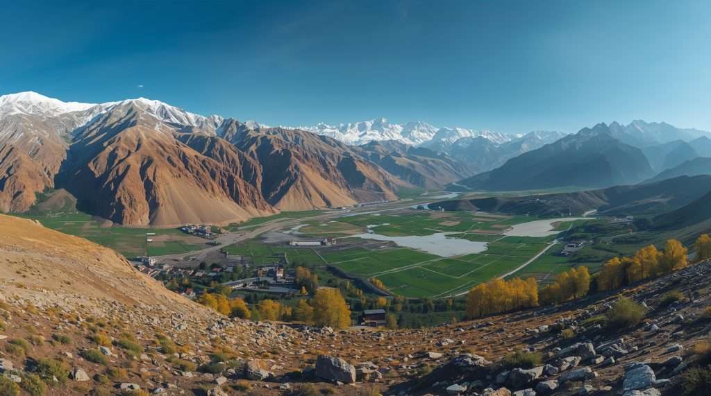 Scenic landscape in Tajikistan featuring a clear river winding through green fields, with majestic, snow-capped mountains rising under a sky dotted with fluffy clouds.