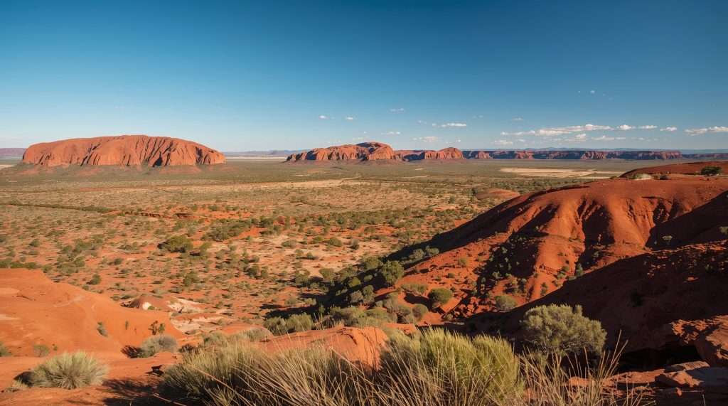 A vast arid landscape in Australia featuring red rock formations and sparse vegetation under a clear blue sky.