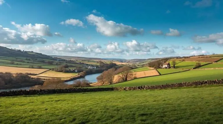 Rolling green fields divided by hedgerows and dry stone walls under a partly cloudy sky, with a river winding through a valley in a rural landscape in the United Kingdom.