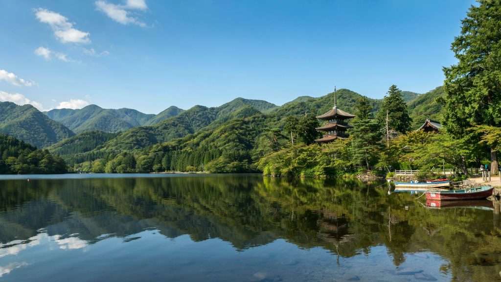 A calm lake reflects green mountains and a pagoda in Japan.